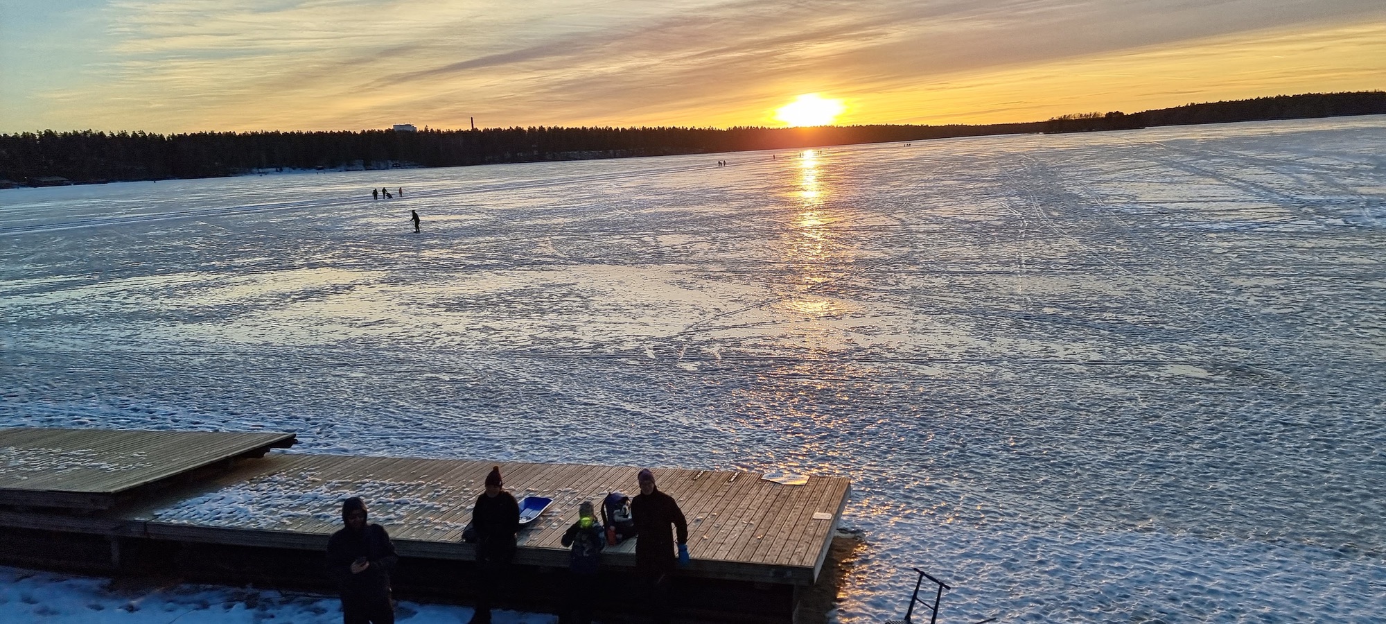 Jäätynyt Sääksjärvi auringonlaskussa. Hiihtäjiä ja kävelijöitä jäällä, laiturilla porukka ja pulkka.